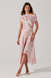 A woman is wearing a sleeveless, off-the-shoulder dress with a pink and white tie-dye pattern. The dress has a fitted waist and a flowing, asymmetrical hemline that is longer in the back and higher in the front, revealing her legs. She is also wearing open-toe heels and gold hoop earrings.
